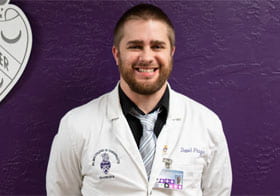 Daniel Prugar, Palmer Florida student, smiling in white clinic coat against a purple background.