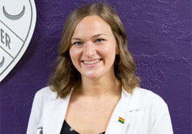 Taylor Taraski smiling in white clinic coat in front of purple background.