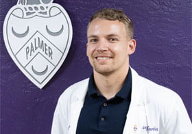 Cory Voorhis smiling in white clinic coat in front of purple background.