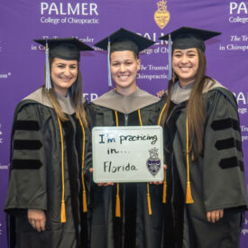 Three students in graduation cap and gowns holding a sign that says "I'm practicing in Florida".