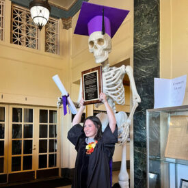 Main Campus graduate in gown posting with skeleton statue.