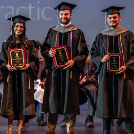 Three graduates in cap and gowns holding awards.