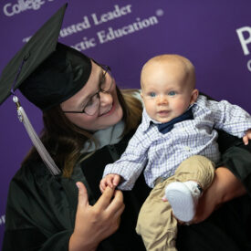 Graduate in cap and gown holding infant.