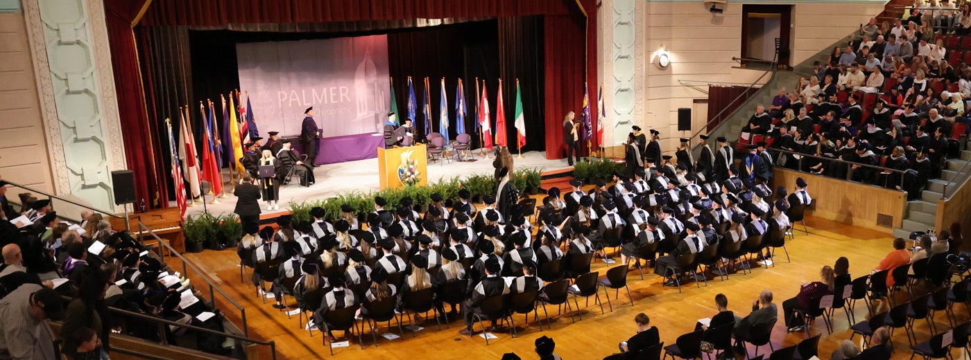 Large group of students sitting with graduation caps and gowns listening to speaker.