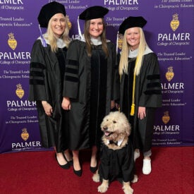 Three students in graduation cap and gowns posing with dog in graduation gown.